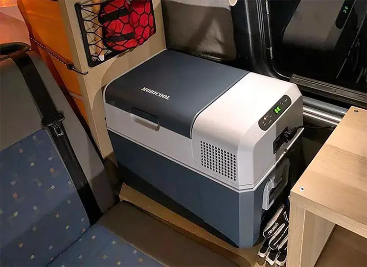 Nevera portátil Mobicool FR40 instalada en una furgoneta camper, mostrando su diseño compacto y eficiente, ideal para viajes y acampadas. La imagen destaca su integración en el espacio interior, con acceso fácil desde los asientos y almacenamiento adicional alrededor, ideal para mantener alimentos y bebidas frías en rutas prolongadas.